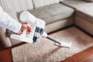 Leistungsstarker Akku-Staubsauger mit weißer Zyklon-Staubsammeltechnologie in der Hand, reinigt den Teppich im Haus in der Nähe des Sofas. foto