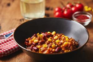 Chili con Carne mit Hackfleisch, Bohnen und Mais in dunkler Schüssel auf Holzhintergrund. mexikanische und texanische küche foto