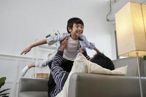 asiatische thailändische familie zusammen, vater legt sich hin und spielt lustig mit dem sohn, indem er hebt, breite die arme wie ein fliegendes flugzeug auf dem sofa im wohnzimmer aus, glückliche freizeiten, schönes wochenende, häuslicher lebensstil des wohlbefindens. foto