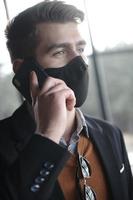 geschäftsmann, der eine medizinische gesichtsmaske des coronavirus trägt, während er das smartphone benutzt foto