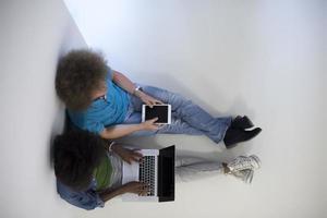 Multiethnisches Paar, das mit einem Laptop und einer Tablet-Draufsicht auf dem Boden sitzt foto