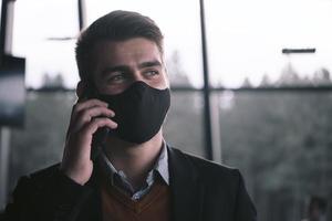 geschäftsmann, der eine medizinische gesichtsmaske des coronavirus trägt, während er das smartphone benutzt foto