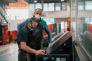 zwei arbeiter, die wegen einer coronavirus-pandemie eine gesichtsmaske tragen, programmieren eine moderne cnc-maschine foto