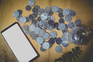 flach gelegte draufsichtmünzen sparen glas und telefon mit isoliertem bildschirm auf holztischsparkonzept foto