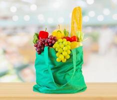frisches obst und gemüse in wiederverwendbarer grüner einkaufstasche auf holztischplatte mit supermarkt-lebensmittelgeschäft unscharfer defokussierter hintergrund mit bokeh-licht foto