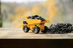 Ein kleiner gelber Spielzeuglastwagen ist mit Sonnenblumenkernen neben einem kleinen Haufen Sonnenblumenkerne beladen. ein Auto auf einer Holzoberfläche vor dem Hintergrund eines herbstlichen Waldes. Transport von Sonnenblumenkernen foto