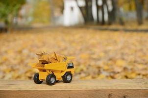 Das Konzept der saisonalen Ernte von Herbstlaub wird in Form eines mit Blättern beladenen gelben Spielzeuglastwagens vor dem Hintergrund des Herbstparks dargestellt foto