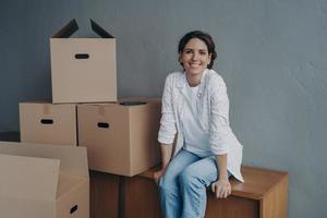 lächelnde hispanische Frau, die mit Kartons in einem neuen Miethaus sitzt. Vermietung von Immobilien, Umzug foto