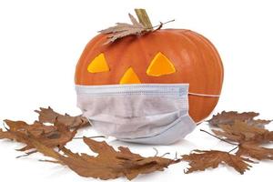 halloween, orangefarbener kürbis mit medizinischer einwegmaske und trockenen herbstblättern. weißer Hintergrund. foto