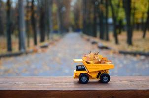 Das Konzept der saisonalen Ernte von Herbstlaub wird in Form eines mit Blättern beladenen gelben Spielzeuglastwagens vor dem Hintergrund des Herbstparks dargestellt foto