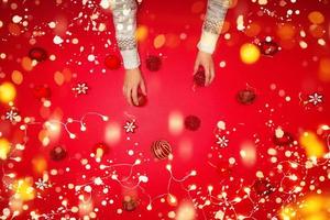 das mädchen benutzt die hand, um die rote kugeldekoration auf rotem hintergrund mit weihnachtsschmuck mit led-licht zu halten. Ansicht von oben. Familientraditionen zu Weihnachten. Konzept Weihnachten. foto