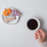 Frauenhand, die Kaffeetasse hält, während sie lustige Halloween-Plätzchen isst. Happy Halloween Day, Trick or Threat, Hallo Oktober, Herbstherbst, Traditions-, Party- und Urlaubskonzept foto