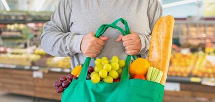 Kunden halten wiederverwendbare grüne Einkaufstasche mit Obst und Gemüse über Supermarktgang verschwimmen defokussierte Produktregale im Inneren von Bokeh hellem Hintergrund foto