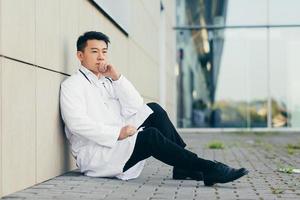 porträt arzt asiatischer mann müde nach der arbeit auf dem boden in der nähe der klinik sitzend enttäuscht foto