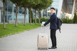 erfolgreicher asiatischer geschäftsmann in der nähe von flughafen und busbahnhof geht mit koffern ernst foto