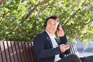 Fröhlicher asiatischer Geschäftsmann, der auf einer Bank in einem Stadtpark in der Innenstadt sitzt und Musik hört foto