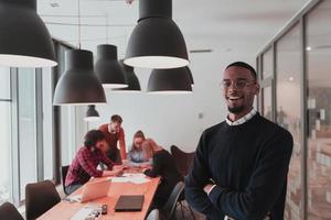 Porträt eines glücklichen, tausendjährigen männlichen Geschäftsinhabers im modernen Büro. Geschäftsmann mit Brille, lächelnd und Blick in die Kamera. beschäftigtes vielfältiges team, das im hintergrund arbeitet. Führungskonzept. Kopfschuss. foto