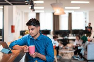 Lässiger Geschäftsmann, der mit dem Laptop eine Pause von der Arbeit einlegt, während er Tee im Entspannungsbereich des modernen Großraum-Startup-Büros trinkt foto