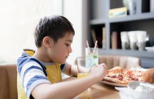 Kind, das hausgemachte Pizza im Café isst, fröhlicher Kinderjunge, der im Restaurant ein großes Stück frisch zubereiteter Pizza abbeißt, Konzept der glücklichen Familienzeit foto