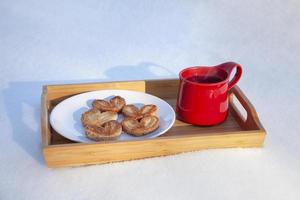 roter Becher mit Keksen auf dem Schnee rote Keramiktasse mit heißem Tee und Blätterteig-Zuckerkeksen in Form von Herzen auf einem Holztablett, auf Schnee an einem sonnigen Tag, unscharfer Hintergrund, Konzept der Liebe, foto