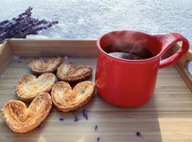 roter Becher mit Keksen auf der schneebedeckten Keramiktasse mit heißem Tee und Blätterteig-Zuckerplätzchen in Form von Herzen auf einem Holztablett, auf Schnee an einem sonnigen Tag, unscharfer Hintergrund, Konzept der Liebe foto