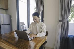 ein junger asiatischer mann verbringt seine zeit zu hause im esszimmer und arbeitet glücklich an seinem laptop foto