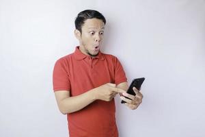 überraschter asiatischer Mann mit rotem T-Shirt, der auf sein Smartphone zeigt, isoliert durch weißen Hintergrund foto