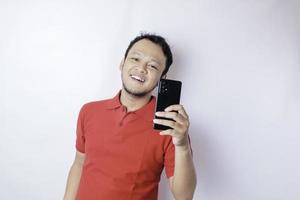 ein porträt eines glücklichen asiatischen mannes lächelt und hält sein smartphone mit einem roten t-shirt, das durch einen weißen hintergrund isoliert ist foto