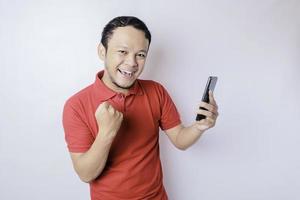 ein junger asiatischer mann mit einem glücklichen erfolgreichen ausdruck, der rotes hemd trägt und smartphone lokalisiert durch weißen hintergrund hält foto
