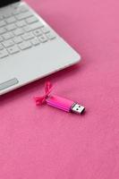 Eine leuchtend rosa USB-Flash-Speicherkarte mit einer rosa Schleife liegt auf einer Decke aus weichem und pelzigem hellrosa Fleece-Stoff neben einem weißen Laptop. klassisches weibliches Geschenkdesign für eine Speicherkarte foto