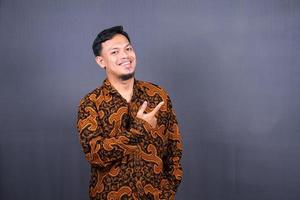 Porträt eines glücklichen jungen Mannes in Batik-Indonesien, der mit dem Finger auf den Kopierraum zeigt, der über grauem Hintergrund isoliert ist foto
