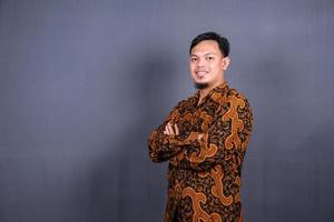 Porträt eines attraktiven jungen asiatischen Mannes in Batik-Indonesien, der mit verschränkten Armen auf grauem Hintergrund steht foto