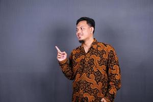 Porträt eines glücklichen jungen Mannes in Batik-Indonesien, der mit dem Finger auf den Kopierraum zeigt, der über grauem Hintergrund isoliert ist foto