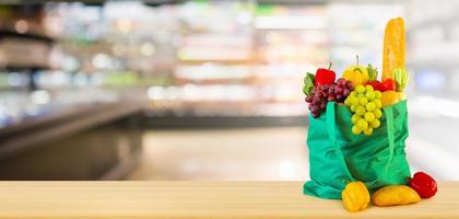 frisches obst und gemüse in wiederverwendbarer grüner einkaufstasche auf holztischplatte mit supermarkt-lebensmittelgeschäft unscharfer defokussierter hintergrund mit bokeh-licht foto