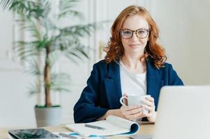 angenehm aussehende, elegante Freiberuflerin bucht Artikel im Webshop, liest Nachrichten im Internet, schreibt Notizen in den Notizblock, arbeitet am Laptop, gekleidet in elegantes Outfit. Geschäfts- und Arbeitskonzept foto