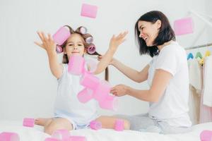 horizontale aufnahme eines schönen kleinen kindes hat lockenwickler auf dem kopf, spielt mit ihnen und wird zu karneval eine wunderbare frisur haben, isoliert über weißem hintergrund. lustige tochter spielt zusammen mit mama foto