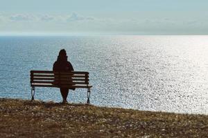 Ein einzelnes Mädchen in einer schwarzen Jacke und einem Hut, das auf einer Bank an einer Klippe am friedlichen, ruhigen Ort vor dem Meer sitzt foto