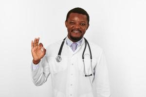 Lächelnder schwarzer bärtiger Arzt in weißer Robe mit Stethoskop zeigt ok Geste, weißer Hintergrund foto