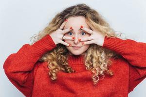 Horizontales Porträt einer mysteriösen blauäugigen Frau mit lockigem hellem Haar, bedeckt das Gesicht mit den Händen, gekleidet in einen lockeren roten Pullover, isoliert auf weißem Hintergrund. überraschung und menschenkonzept foto