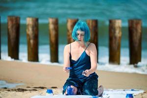 Künstlerische blauhaarige Performance-Künstlerin mit blauen Gouachefarben, die am Strand sitzen foto