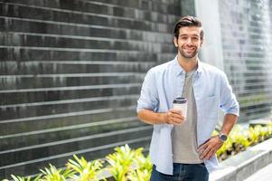 junger kaukasischer mann, der kaffee trinkt und ein lässiges hemd trägt, während er auf seinem weg geht, um sich in seinem urlaub zu entspannen. foto