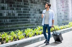 junger kaukasischer mann, der kaffee trinkt und den koffer zieht und auf seinen freund wartet, um gemeinsam zu reisen. foto