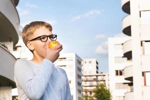 Süßer Schuljunge mit Brille, der an einem sonnigen Tag Apfel gegen den blauen Himmel isst. foto