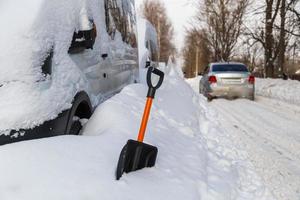 Kunststoff-Schneeschaufel vor schneebedecktem Minivan am sonnigen Wintermorgen und silbernes Auto, das auf verschwommenem Hintergrund vorbeifährt foto