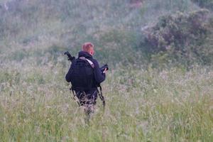 Profi-Fotograf bewegt sich am nebligen Morgen durch hohes trockenes Gras zum Aufnahmeort mit Kamera, Stativ und schwarzem Rucksack foto