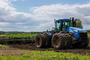 Blauer Traktor mit Doppelrädern, der an heißen sonnigen Tagen eine Scheibenegge mit Rollenkorb zieht foto