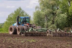 Blauer Traktor mit Doppelrädern, der an heißen sonnigen Tagen eine Scheibenegge mit Rollenkorb zieht foto