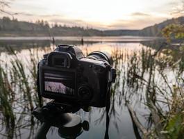 Schwarze Digitalkamera auf Stativ, die frühnebelige Morgenlandschaft am Herbstsee mit selektivem Fokus aufnimmt foto