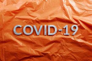 das Wort covid-19 mit Aluminiumbuchstaben auf zerknittertem orangefarbenem Kunststofffolienhintergrund in flacher Laienzusammensetzung, direkt über der Ansicht foto