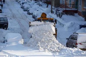 traktor, der am wintertag schnee vom parkplatz in der nähe des wohngebäudes entfernt